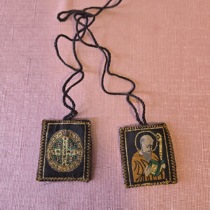 Brown embroidered fabric scapular. The front features a half-length image of Saint Benedict, and the back features the cross of Saint Benedict.   Scapular badge measures 1.35 inches wide, 1.45 inches high. Scapular wire length: panel to panel: 14.5 inches. Escapulario de tela color café bordado  En la parte delantera muestra imagen de San Benito de medio cuerpo,  en la parte de atrás la cruz de San Benito Medidas de la insignia escapular 1.35 pulgadas ancho  de la insignia escapular, 1.45 pulgadas alto Longitud del cable escapular: panel a panel 14.5 pulgadas.
