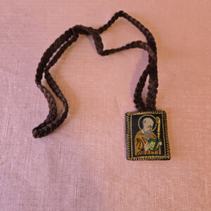 Brown embroidered fabric braided scapular. The front features a half-length image of Saint Benedict, and the back features the cross of Saint Benedict. Scapular badge measures 1.35 inches wide, 1.45 inches high. Scapular wire length: panel to panel: 14.5 inches. Escapulario de tela color café bordado  En la parte delantera muestra imagen de San Benito de medio cuerpo,  en la parte de atrás la cruz de San Benito Medidas de la insignia escapular 1.35 pulgadas ancho  de la insignia escapular, 1.45 pulgadas alto Longitud del cable escapular: panel a panel 14.5 pulgadas.